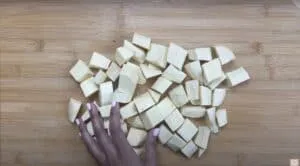 Chopped raw plantains on a wooden cutting board, hand arranging pieces, ready for cooking.